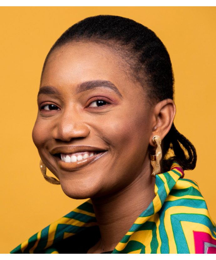 Dee, smiling, photographed against a yellow backdrop in a green and yellow wax-print top