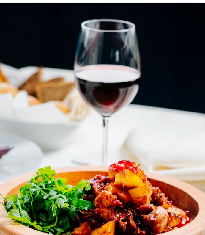 A bowl of pepper stew with rocket beside a glass of red wine on a laid table